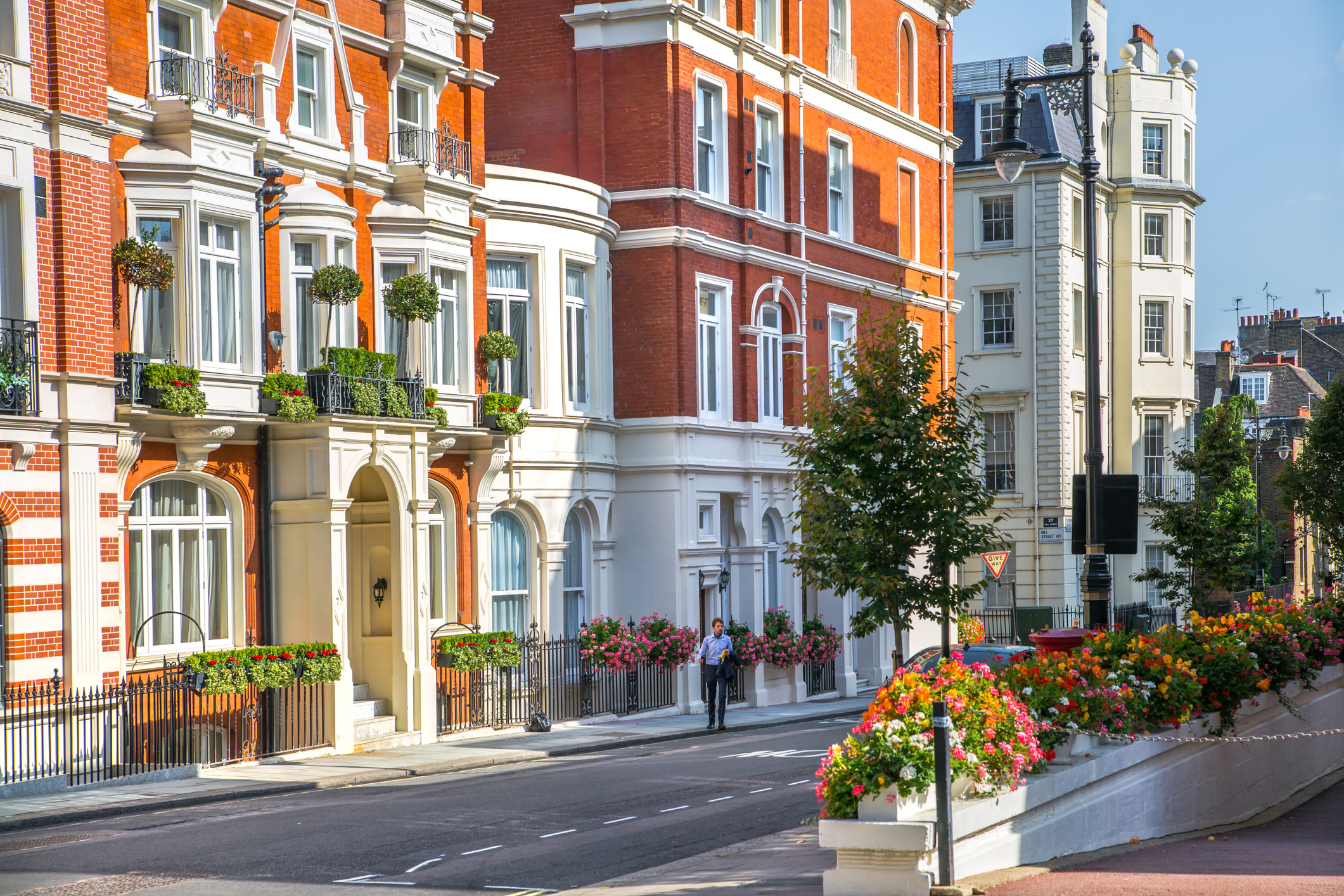Row of elegant red-brick and white London townhouses with bay windows, balconies and flower boxes on a sunny day, man walking along quiet street.