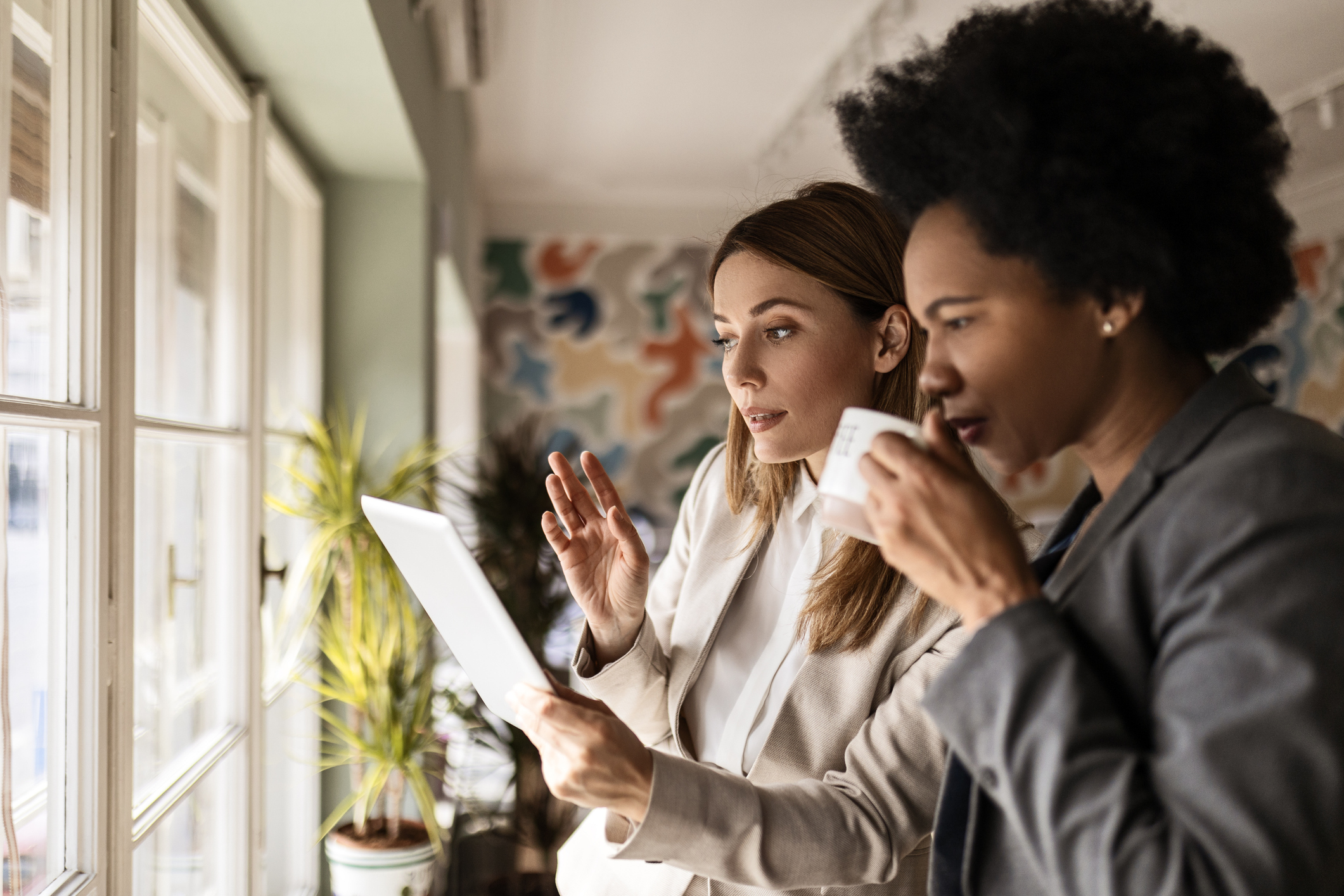 Two business women standing by a large window in a modern office, discussing the property strategy.