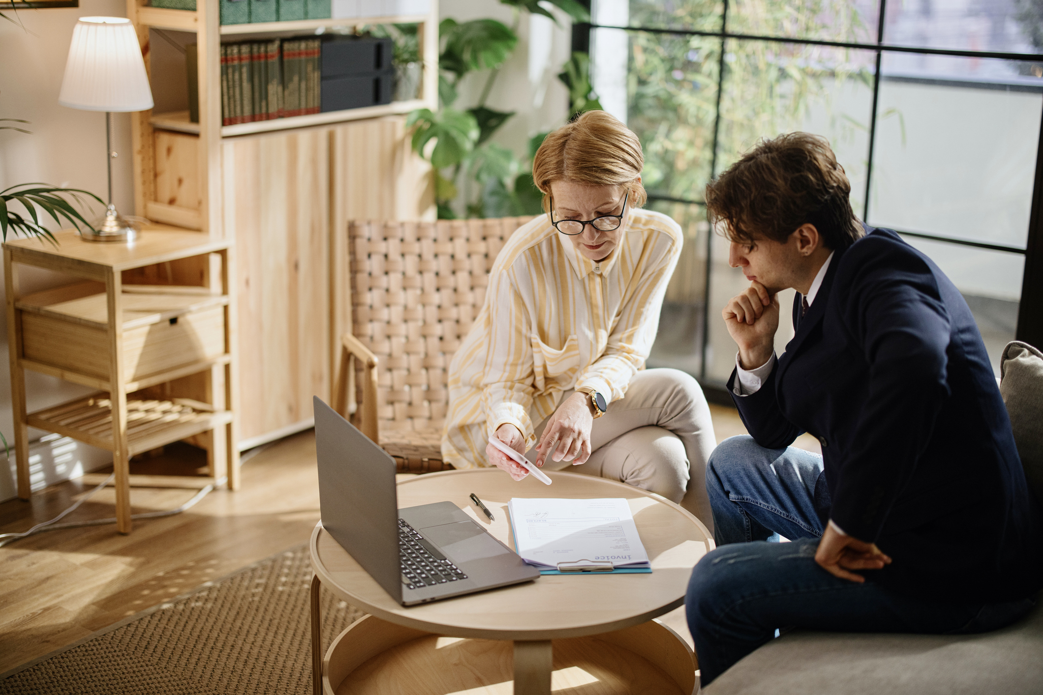 Two people in a modern office lounge sitting at a round coffee table, discussing documents and a laptop during an investment coaching meeting.