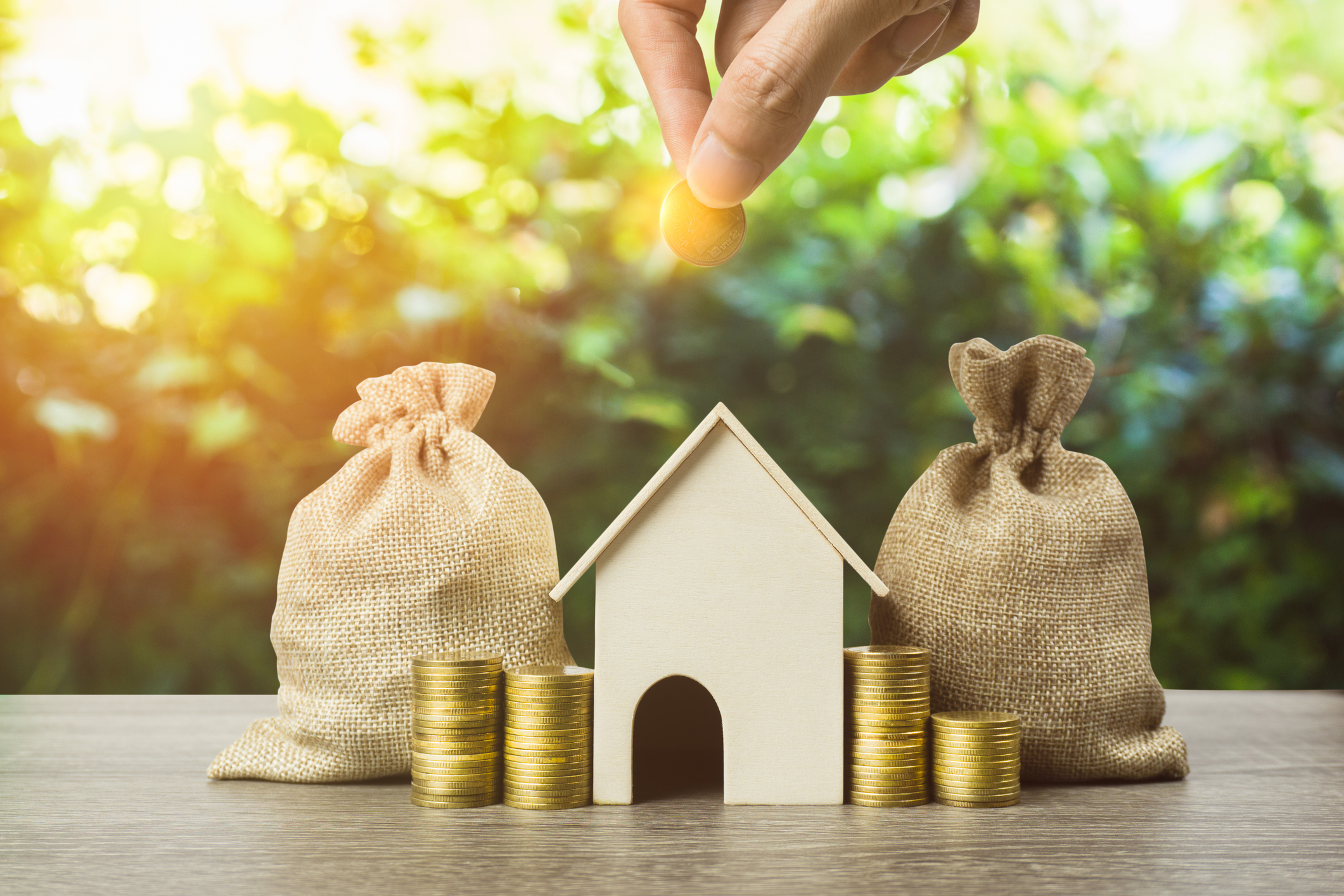 Hand placing a coin above a small wooden house between money bags and coin stacks, symbolising property investment and savings.