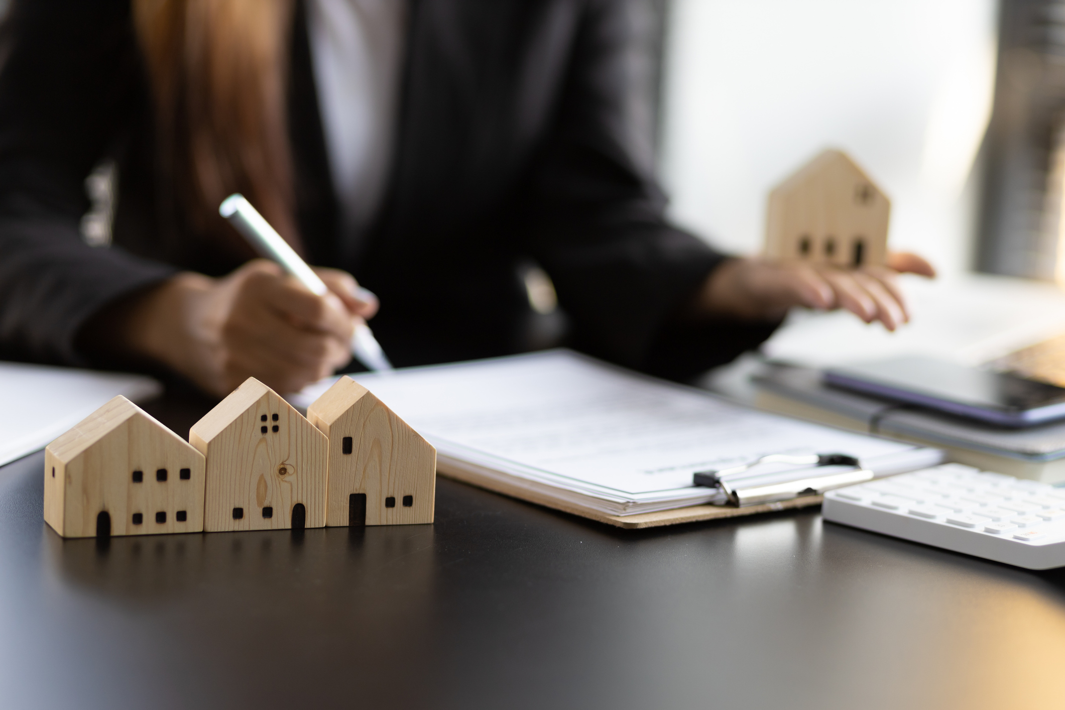 A person signing property investment paperwork at a desk with wooden house models, clipboard and calculator.