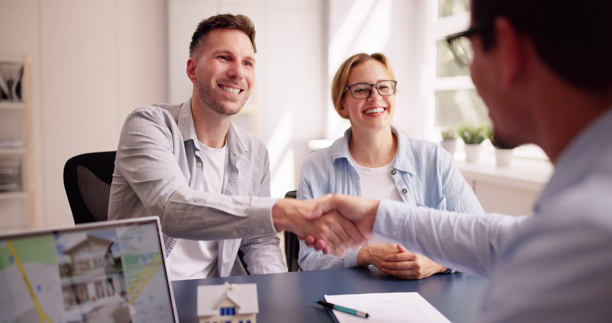 Smiling couple shake hands with a property adviser after agreeing a deal, with house model and paperwork on the desk.