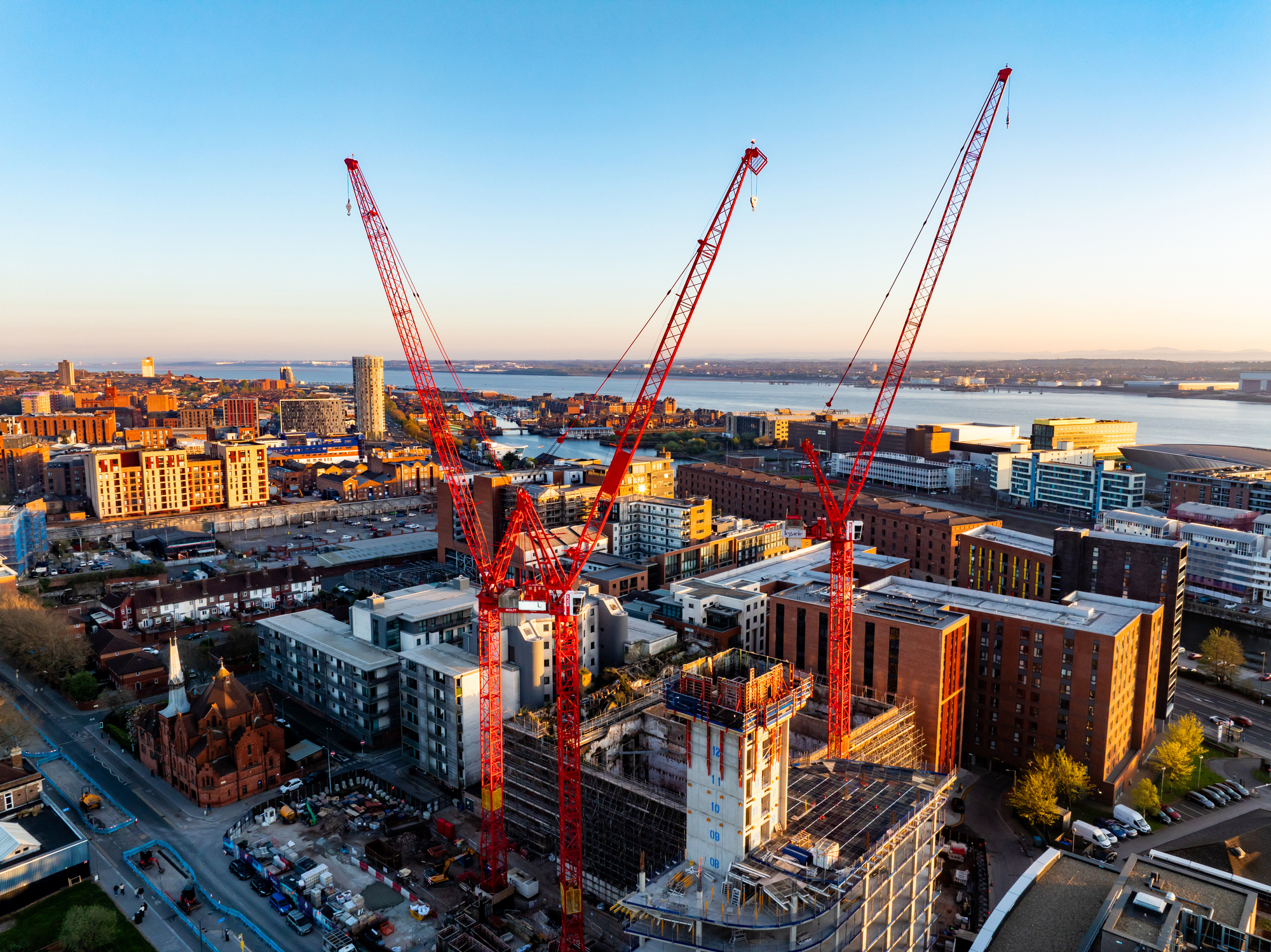 Aerial view of a modern city waterfront with several bright red construction cranes towering above new high-rise buildings at sunrise.